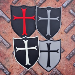 Op de afbeelding: Vier geborduurde patches in de vorm van een schild met een kruis erin. De patches zijn zwart, grijs en wit met rode, grijze en witte kruisen.