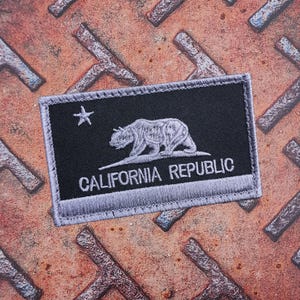 Peut inclure: Un écusson rectangulaire représentant le drapeau de l'État de Californie. L'écusson a un fond noir avec un ours blanc, une étoile et les mots "CALIFORNIA REPUBLIC" en blanc. La bordure est grise.