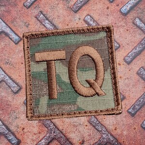 May include: Square patch with a camouflage pattern background and the embroidered letters "TQ" in brown. The patch has a brown border and is set against a rusty metal surface.
