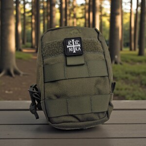 Pode incluir: Bolsa t&aacute;tica verde-oliva com v&aacute;rias correias e um patch de velcro. O patch apresenta uma cruz branca e o texto "IC XC NIKA". A bolsa &eacute; feita de tecido dur&aacute;vel e tem um pequeno la&ccedil;o na lateral.