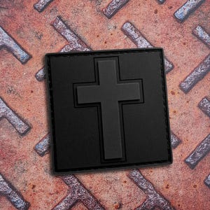 Pode incluir: Um emblema quadrado preto com uma cruz tridimensional em relevo centrada na sua superfície. O emblema tem um acabamento mate liso com bordas costuradas, apresentando um design limpo e uniforme que se destaca contra o fundo texturizado.