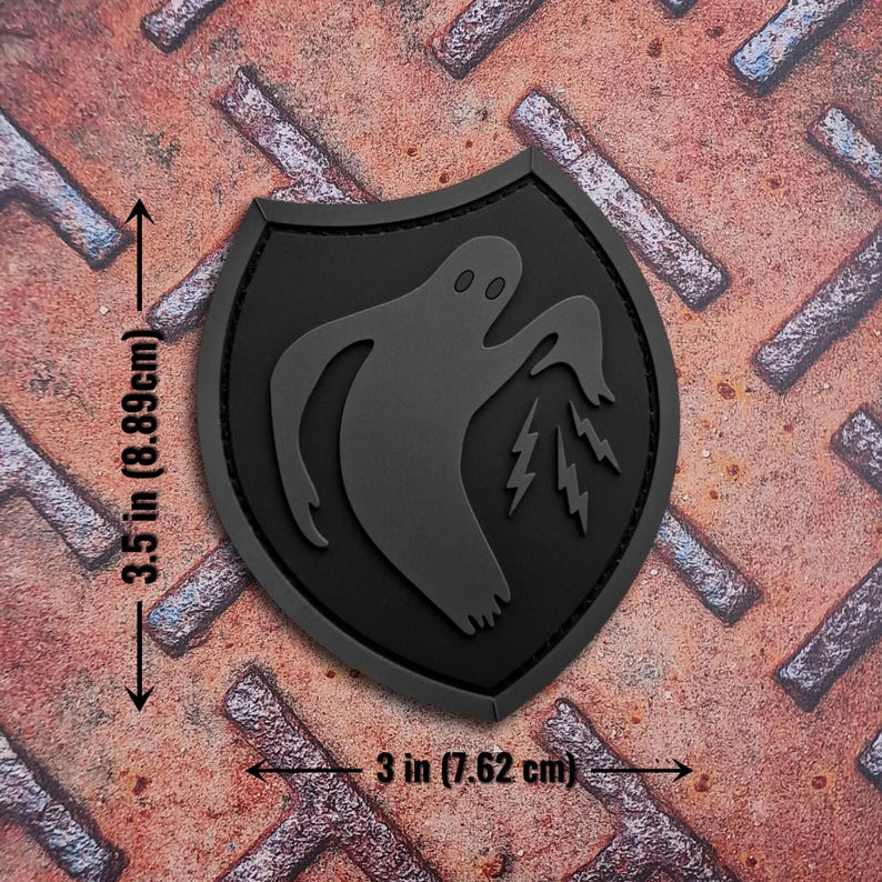 Op de afbeelding: Zwart, schildvormig embleem met een spook- en bliksemontwerp. Het embleem is 8,89 cm hoog en 7,62 cm breed. Gemaakt van een rubberachtig materiaal, waarschijnlijk een embleem of patch voor kleding of accessoires.