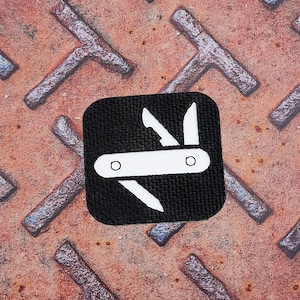 Può includere: Una toppa quadrata nera con un'illustrazione bianca di un coltello tascabile, con lama e forbici. La toppa è posta su uno sfondo metallico arrugginito e strutturato con un motivo ripetuto di elementi a forma di T.