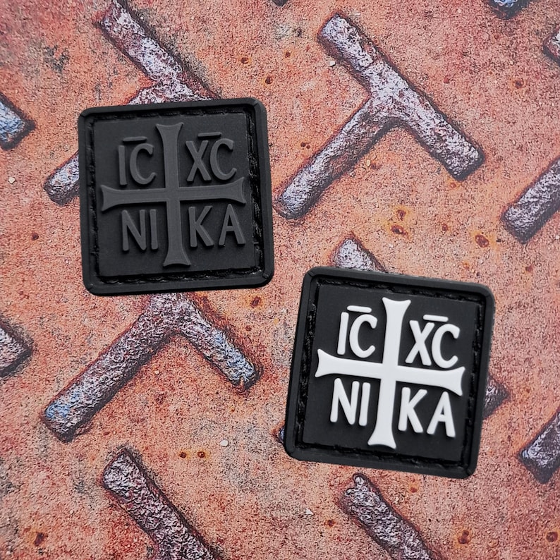 Pode incluir: Duas aplica&ccedil;&otilde;es quadradas pretas com um desenho de cruz e o texto "IC XC NIKA". Uma aplica&ccedil;&atilde;o tem uma cruz preta sobre fundo preto, a outra uma cruz branca sobre preto. Est&atilde;o sobre uma superf&iacute;cie met&aacute;lica enferrujada e texturizada.