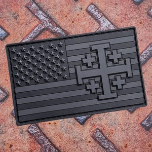 Op de afbeelding: Een rechthoekige patch met een gestileerd ontwerp in grijstinten. De linkerkant toont de sterren en strepen van de Amerikaanse vlag. De rechterkant toont een groot Jeruzalemkruis.
