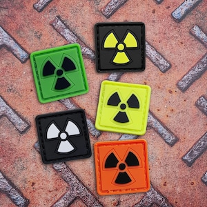 Pode incluir: Cinco patches quadrados com um símbolo de radiação em várias combinações de cores: verde com preto, preto com amarelo, amarelo com preto, preto com branco e laranja com preto. Os patches estão sobre uma superfície metálica enferrujada e texturizada.