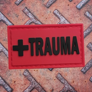 May include: A red rectangular patch with the word "+TRAUMA" in black, block letters. A black cross is on the left side of the text. The patch is on a rusty, textured metal surface.