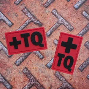 May include: Two red rectangular patches with black text and a black cross. The text on the patches reads "+TQ". The patches are on a rusty metal surface with a repeating pattern.