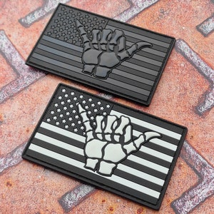 Pode incluir: Dois patches bordados em preto e branco com um design de bandeira americana e uma mão de esqueleto fazendo o sinal shaka. Os patches estão sobre uma superfície marrom texturizada.