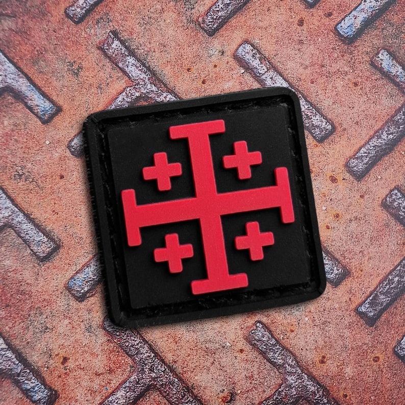 May include: A square black patch featuring a red Jerusalem cross design. The cross has a large central cross with four smaller crosses in each quadrant. The patch is set against a textured, rusty metal surface.