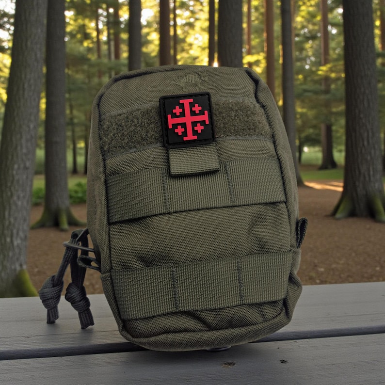 May include: An olive green tactical pouch with a red and black patch featuring a Jerusalem cross. The pouch has multiple straps and a small loop with a cord. The background is a blurred forest scene.