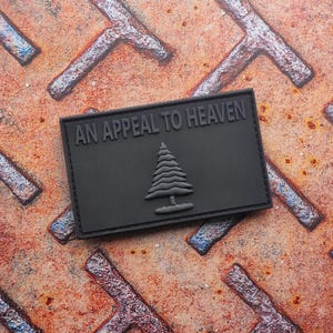 Może przedstawiać: Czarna prostokątna naszywka z wytłoczonym napisem "AN APPEAL TO HEAVEN" u góry. Poniżej znajduje się stylizowane drzewo. Naszywka jest umieszczona na teksturowanej, zardzewiałej metalowej powierzchni.