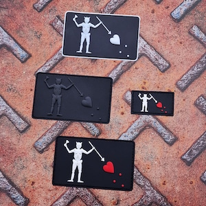 Op de afbeelding: Vier geborduurde patches in zwart en wit met een skelet dat een zwaard en een hart vasthoudt. De patches zijn op een roestige metalen achtergrond.