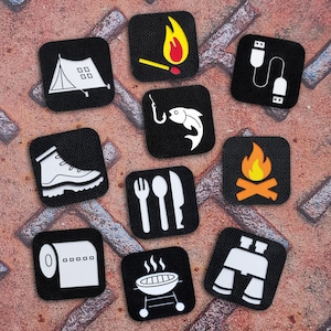 May include: A collection of black square patches with white and orange camping-themed icons. Icons include a tent, a match with a flame, a USB cable, a hiking boot, a fish on a hook, a fork, spoon, and knife, a campfire, a roll of paper towels, a barbecue grill, and binoculars.