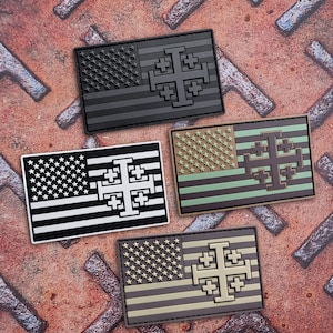 May include: Four rectangular patches featuring the American flag design with a Jerusalem cross. The patches are in various color schemes: black, black and white, olive green and brown, and brown and tan. The patches are on a textured, rusty metal surface.