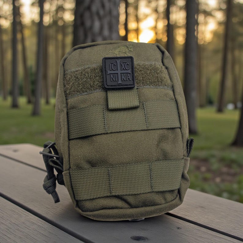 Pode incluir: Bolsa t&aacute;tica verde-oliva com v&aacute;rias correias e um patch preto com texto branco. A bolsa &eacute; retangular com bordas arredondadas e uma pequena presilha na lateral. O fundo &eacute; uma cena de floresta desfocada.