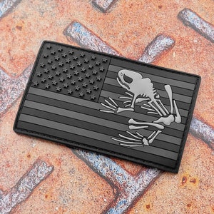 May include: A black and gray patch featuring the American flag with a frog skeleton overlay. The flag design includes stars and stripes, with the frog skeleton in a light gray color. The patch is rectangular and has a textured appearance.