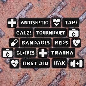 Op de afbeelding: Een verzameling zwarte rechthoekige patches met witte tekst en iconen die medische benodigdheden vertegenwoordigen. De labels bevatten Antiseptic, Gauze, Tourniquet, Tape, Bandages, Meds, Gloves, Trauma, First Aid en IFAK, naast medische symbolen.