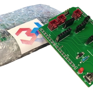 Può includere: Una scheda di circuito verde con connettori neri e rossi, un piccolo pulsante e il testo "R3" stampato su di essa. La scheda è confezionata in un sacchetto di plastica trasparente con un logo.
