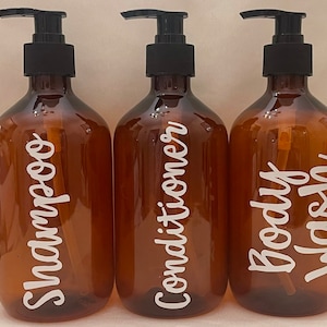 Puede incluir: Tres botellas de vidrio ámbar con tapas de bomba negras y etiquetas blancas. Las etiquetas dicen "Champú", "Acondicionador" y "Gel de baño".