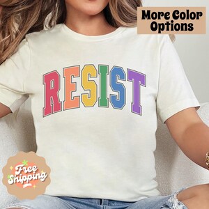 May include: A white t-shirt with the word "RESIST" printed in rainbow colors. The letters are outlined in black and have a varsity style font.