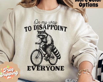 May include: A beige sweatshirt with the text "On my way to disappoint everyone" and a black and white illustration of a raccoon riding a bicycle.