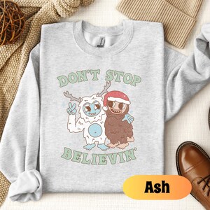 May include: A gray sweatshirt with a white yeti and a brown yeti wearing a Santa hat. The text "Don't Stop Believin'" is printed on the sweatshirt. The sweatshirt is available in the color Ash.