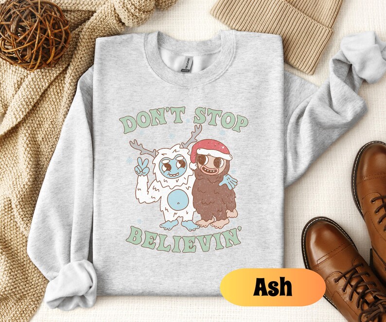 May include: A gray sweatshirt with a white yeti and a brown yeti wearing a Santa hat. The text "Don't Stop Believin'" is printed on the sweatshirt. The sweatshirt is available in the color Ash.