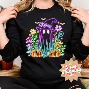 K&ouml;nnte beinhalten: Schwarzer Pullover mit einem neon-gr&uuml;nen, violetten und rosafarbenen Grafikdruck eines Geistes, der einen Hexenhut tr&auml;gt und von Blumen und K&uuml;rbissen umgeben ist.
