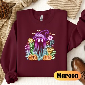 K&ouml;nnte beinhalten: Ein maroonfarbener Pullover mit einem neonpinken und violetten Geist, der einen Hexenhut tr&auml;gt und einen K&uuml;rbis h&auml;lt. Der Geist ist von Blumen und K&uuml;rbissen umgeben. Der Text "Maroon" ist in der rechten unteren Ecke des Pullovers gedruckt.