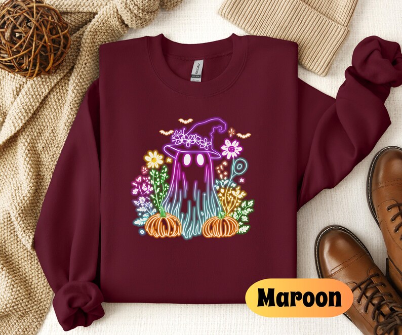 K&ouml;nnte beinhalten: Ein maroonfarbener Pullover mit einem neonpinken und violetten Geist, der einen Hexenhut tr&auml;gt und einen K&uuml;rbis h&auml;lt. Der Geist ist von Blumen und K&uuml;rbissen umgeben. Der Text "Maroon" ist in der rechten unteren Ecke des Pullovers gedruckt.
