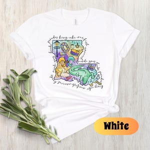 Op de afbeelding: Wit T-shirt met een kleurrijke illustratie van de staat Louisiana. De illustratie bevat een fleur-de-lis, een koningentaart, een paradewagen, een Mardi Gras-masker en de woorden "parades, koningentaart, kreeft, kralen, mardi gras, drankjes, feest".