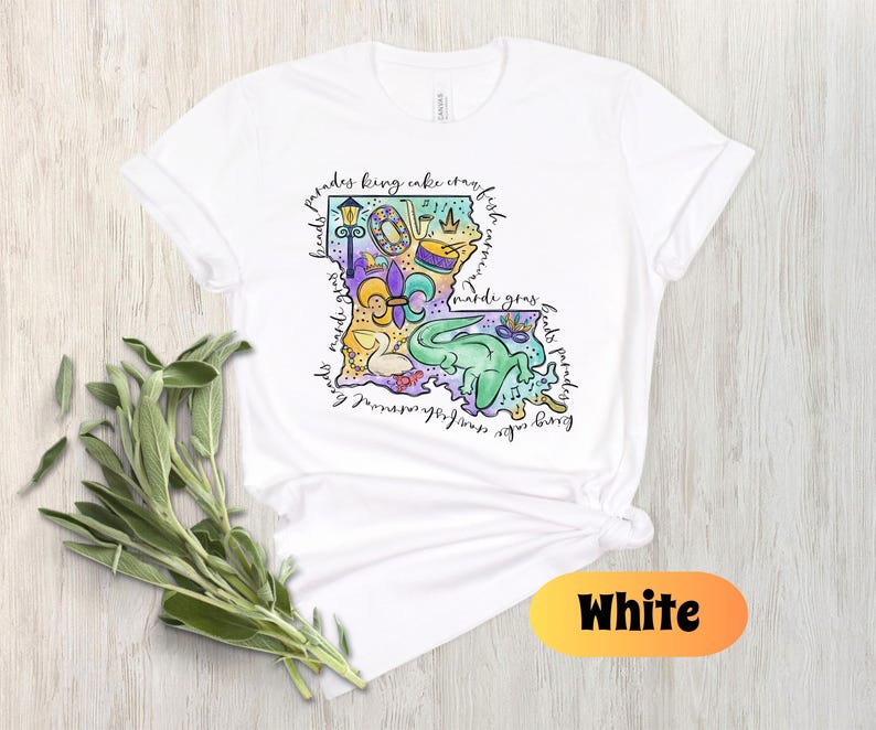 Op de afbeelding: Wit T-shirt met een kleurrijke illustratie van de staat Louisiana. De illustratie bevat een fleur-de-lis, een koningentaart, een paradewagen, een Mardi Gras-masker en de woorden "parades, koningentaart, kreeft, kralen, mardi gras, drankjes, feest".