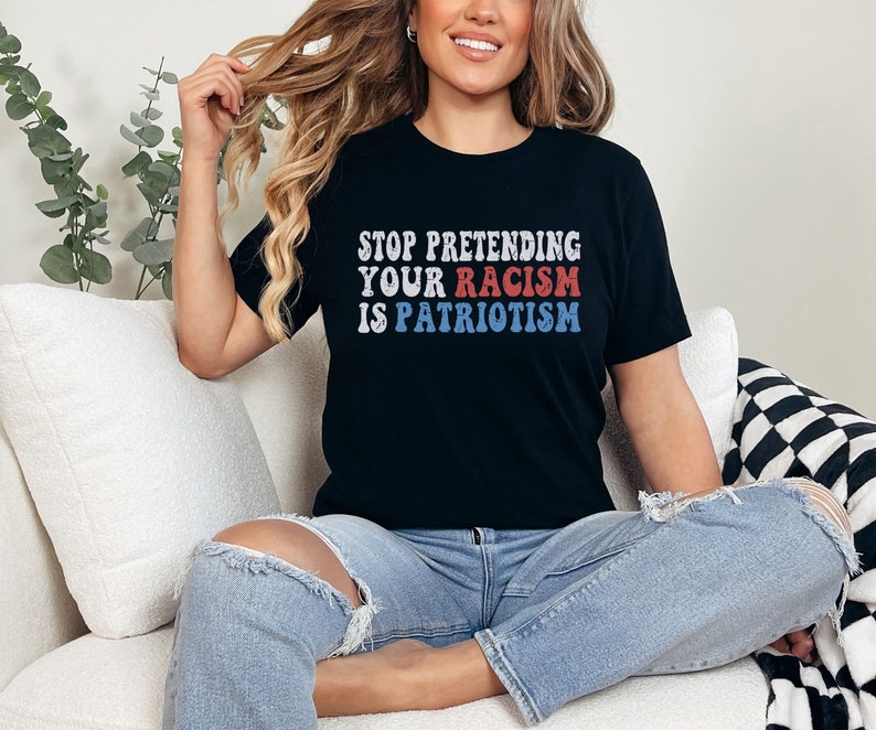 May include: A black t-shirt with the text "STOP PRETENDING YOUR RACISM IS PATRIOTISM" in red, white, and blue letters. The shirt is being worn by a person sitting on a white couch.