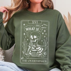 Puede incluir: Sudadera verde oliva con un dise&ntilde;o blanco de un esqueleto sosteniendo una taza, con el texto "WHAT IF..." y "THE OVERTHINKER". El dise&ntilde;o se asemeja a una carta del tarot, con detalles florales y estrellas.