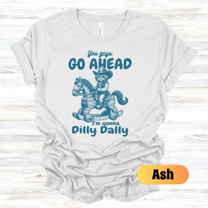 May include: A light grey t-shirt with a blue graphic of a cat riding a rocking horse. The text "You guys go ahead I'm gonna dilly dally" is printed on the shirt. The shirt is available in the color Ash.