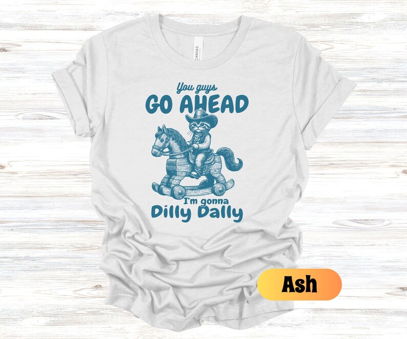 May include: A light grey t-shirt with a blue graphic of a cat riding a rocking horse. The text "You guys go ahead I'm gonna dilly dally" is printed on the shirt. The shirt is available in the color Ash.