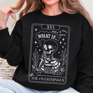 Puede incluir: Sudadera negra con un dise&ntilde;o de carta de tarot blanco. El gr&aacute;fico muestra un esqueleto sosteniendo una taza, con el texto "WHAT IF..." y "THE OVERTHINKER". El dise&ntilde;o incluye estrellas, hojas y el n&uacute;mero romano XVI.