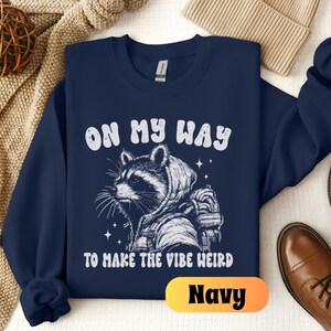 May include: Navy blue sweatshirt with a white graphic of a raccoon wearing a space suit and the text "On My Way To Make The Vibe Weird".