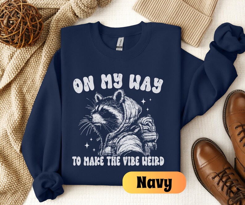 May include: Navy blue sweatshirt with a white graphic of a raccoon wearing a space suit and the text "On My Way To Make The Vibe Weird".