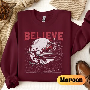 May include: A maroon sweatshirt with the word "BELIEVE" printed in white on the front. The design features a cartoon Santa Claus flying in a spaceship with reindeer pulling it.