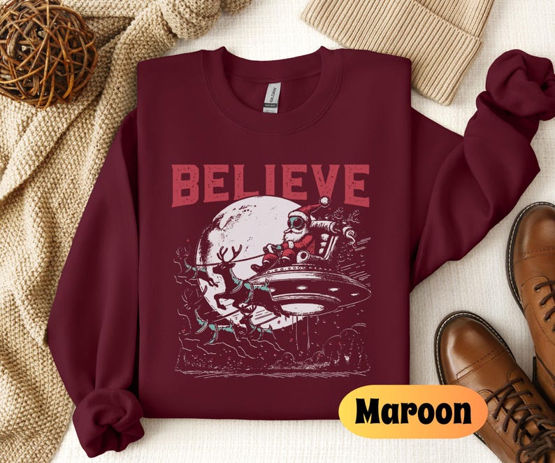 May include: A maroon sweatshirt with the word "BELIEVE" printed in white on the front. The design features a cartoon Santa Claus flying in a spaceship with reindeer pulling it.