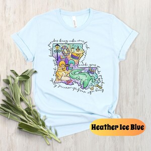 Op de afbeelding: Lichtblauw T-shirt met een kleurrijke illustratie van de staat Louisiana. De illustratie bevat Mardi Gras-parels, een koningentaart, een fleur-de-lis en andere symbolen van de Louisiana-cultuur. De tekst "parades king cake crawfish born in new orleans mardi gras floats parades" omringt de illustratie.