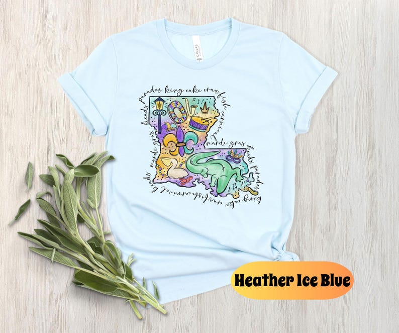 Op de afbeelding: Lichtblauw T-shirt met een kleurrijke illustratie van de staat Louisiana. De illustratie bevat Mardi Gras-parels, een koningentaart, een fleur-de-lis en andere symbolen van de Louisiana-cultuur. De tekst "parades king cake crawfish born in new orleans mardi gras floats parades" omringt de illustratie.