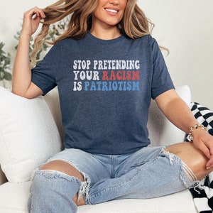 May include: A woman wearing a navy blue t-shirt with the text "STOP PRETENDING YOUR RACISM IS PATRIOTISM" in red, white, and blue letters. She is sitting on a white couch with a black and white checkered throw blanket.