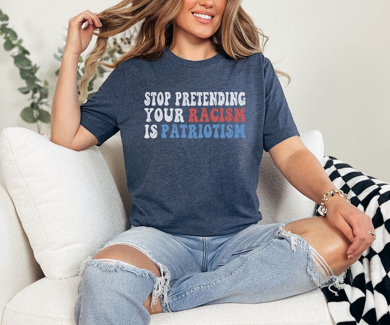 May include: A woman wearing a navy blue t-shirt with the text "STOP PRETENDING YOUR RACISM IS PATRIOTISM" in red, white, and blue letters. She is sitting on a white couch with a black and white checkered throw blanket.