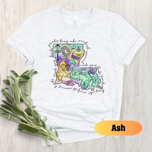 Op de afbeelding: Asgrijze T-shirt met een kleurrijke illustratie van de staat Louisiana met Mardi Gras-thema's en de tekst "parades king cake crawfish beads mardi gras floats parades".
