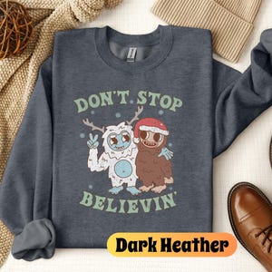 May include: Dark heather grey sweatshirt with a graphic of a yeti and a sasquatch holding hands. The text "Don't Stop Believin'" is printed below the graphic.
