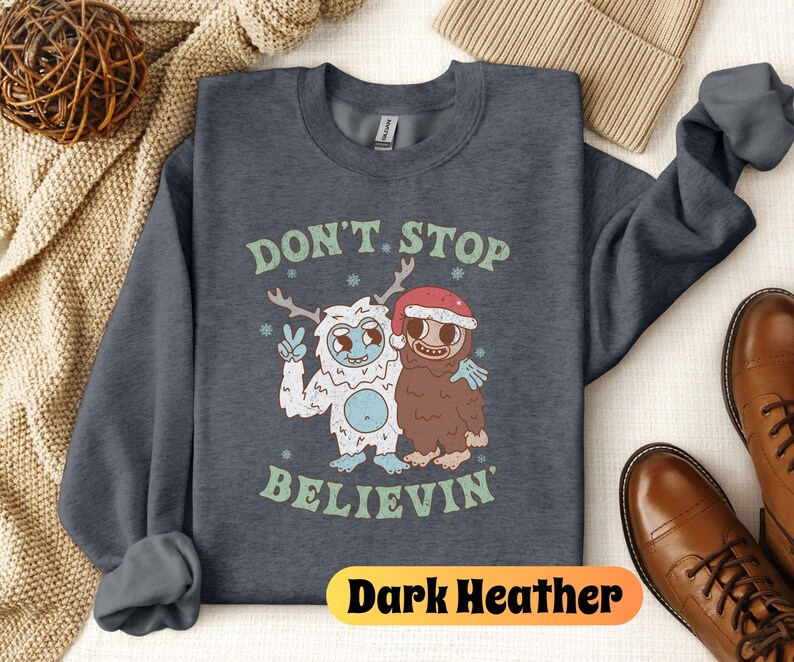 May include: Dark heather grey sweatshirt with a graphic of a yeti and a sasquatch holding hands. The text "Don't Stop Believin'" is printed below the graphic.