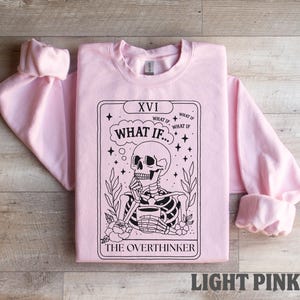 Puede incluir: Sudadera rosa claro con un dise&ntilde;o de carta de tarot en blanco y negro. La carta presenta un esqueleto sosteniendo una taza, con el texto "WHAT IF" y "THE OVERTHINKER". Las mangas est&aacute;n dobladas.